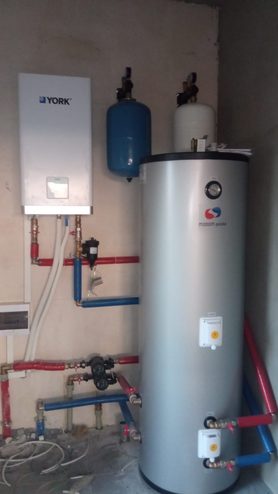 Biały piec gazowy York zawieszony na ścianie, obok pionowy, srebrny zbiornik z logo Modom Polska oraz niebieski zbiornik ciśnieniowy, połączone rurami w kolorach czerwonym i niebieskim.