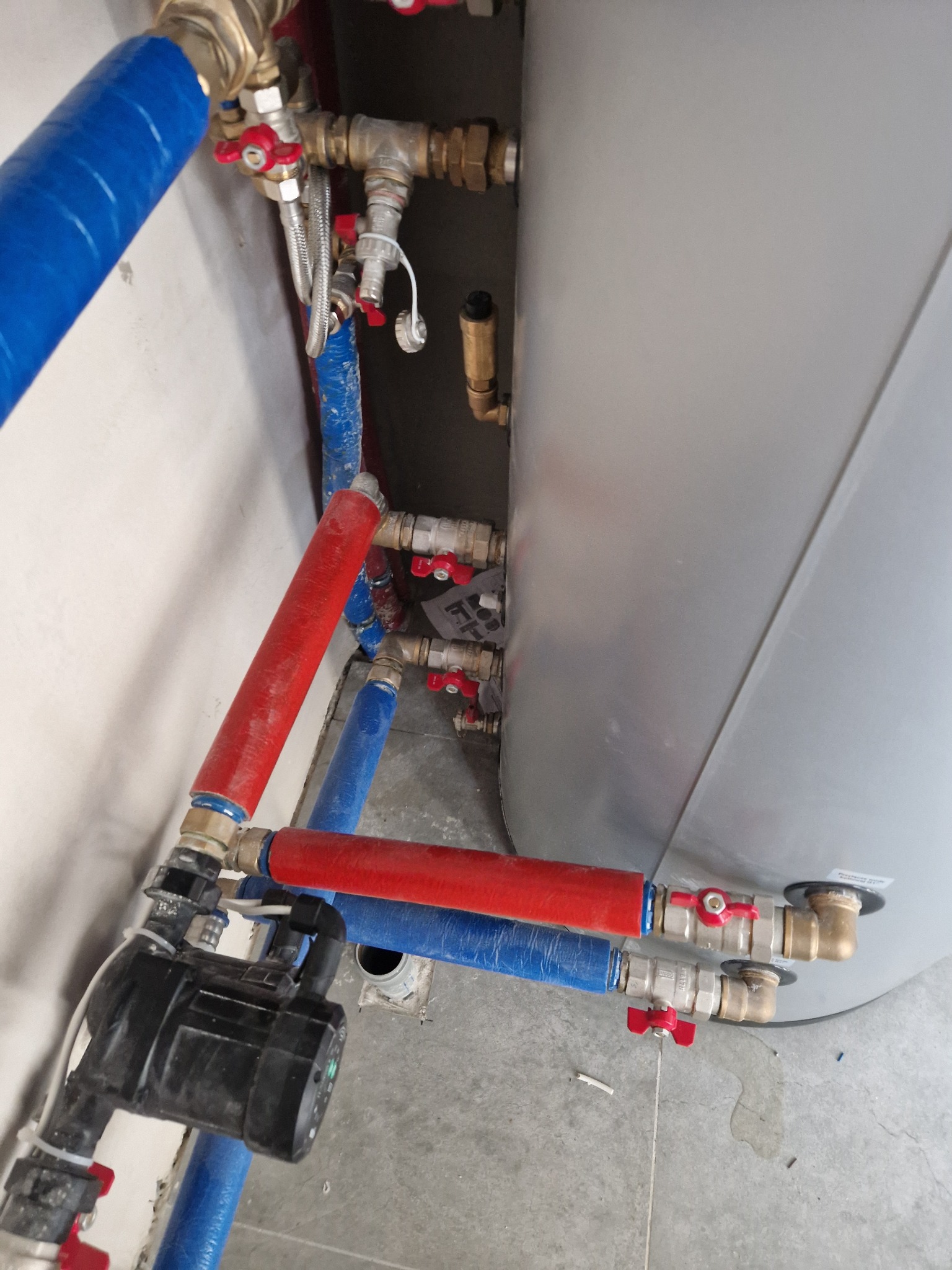 Szare zasobnik ciepłej wody z podłączonymi rurami w kolorze niebieskim i czerwonym, z zaworami z czerwonymi uchwytami, oraz czarna pompa obiegowa na tle szarej ściany i podłogi.