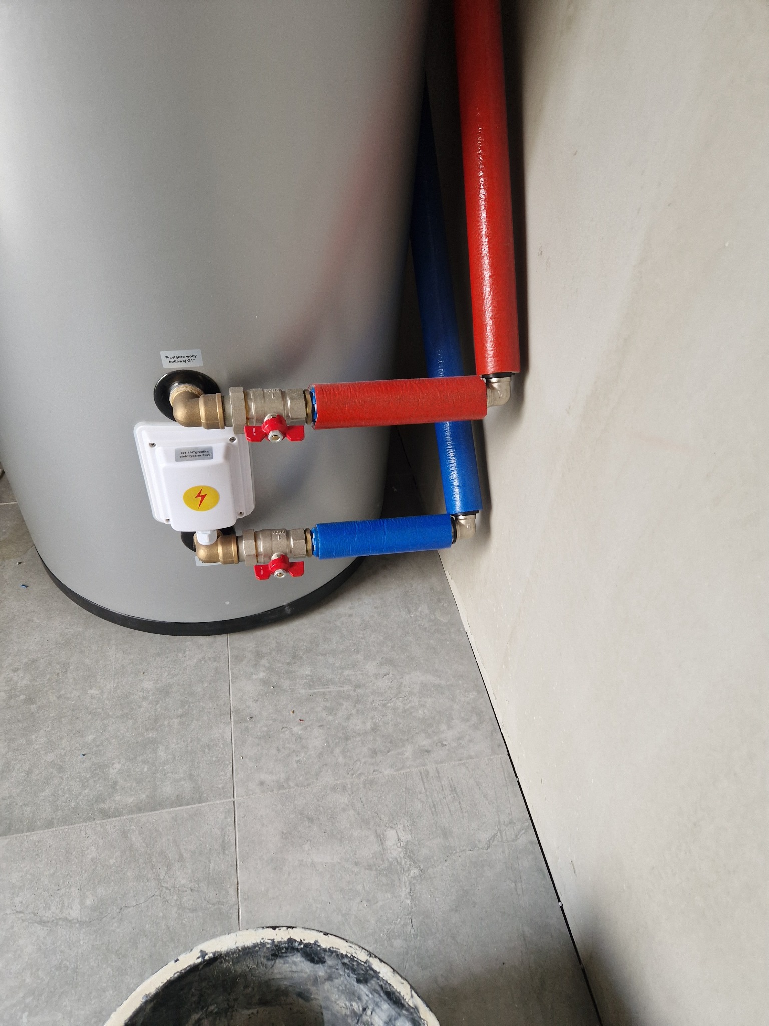 Szary zbiornik z podłączonymi rurami w izolacji czerwonej i niebieskiej, zawory z czerwonymi pokrętłami, biała skrzynka z symbolem błyskawicy, widoczna również podłoga z szarych płytek i fragment...