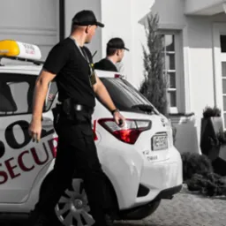 Patrol interwencyjny w czarnych uniformach przy białym samochodzie z logo firmy ochroniarskiej, przed domem z dziećmi w drzwiach.