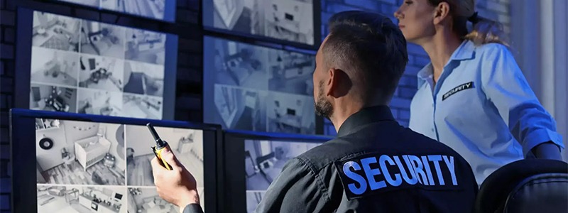 Dwóch pracowników ochrony wpatruje się w ekrany monitoringu, na których widać pokoje dziecięce i inne pomieszczenia, jeden z nich trzyma radiotelefon.