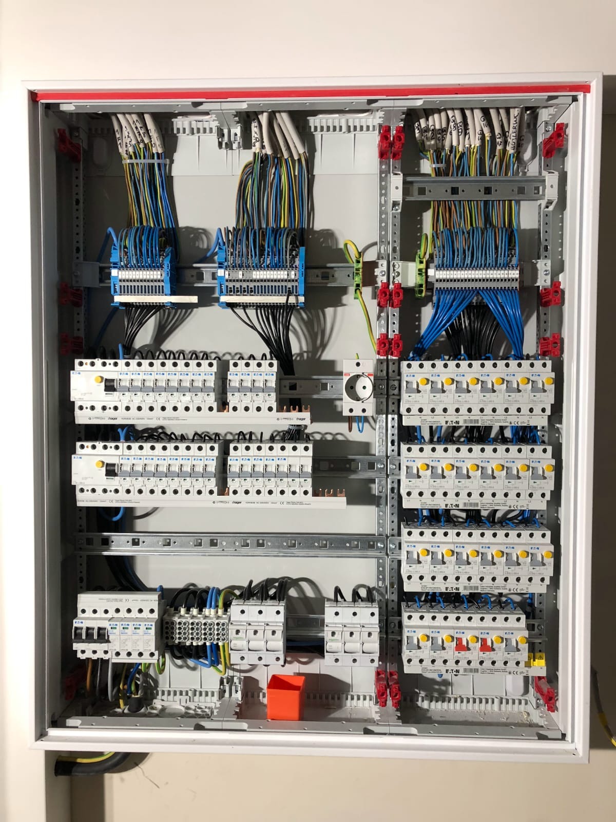 Rozdzielnica elektryczna z widocznymi bezpiecznikami, przewodami oznaczonymi numerami i elementami montażowymi, umieszczona w białej obudowie z czerwoną ramką.