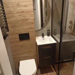Nowoczesna łazienka z drewnopodobnymi płytkami na ścianie, wiszącą toaletą, umywalką z szafką i kabiną prysznicową z czarnymi profilami.