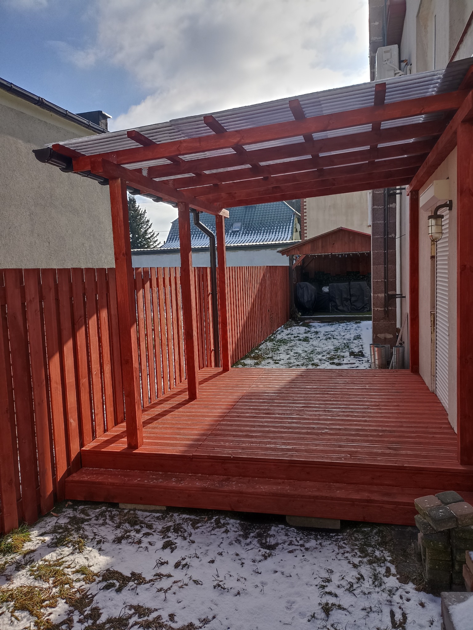 Zadaszony taras z drewnianą podłogą i ogrodzeniem w kolorze czerwieni, częściowo pokryty śniegiem, widok w słoneczny dzień.