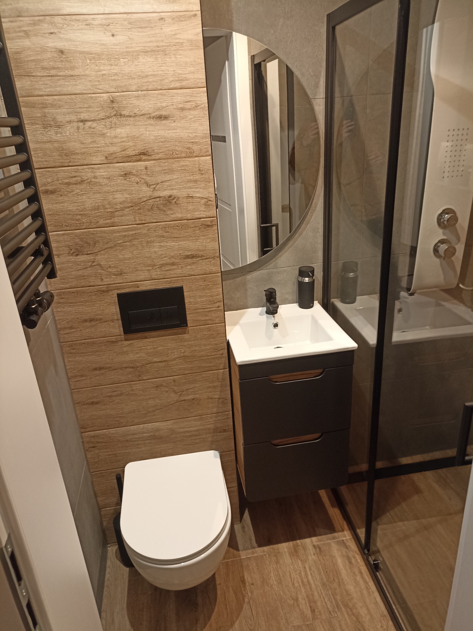 Nowoczesna łazienka z drewnopodobnymi płytkami na ścianie, wiszącą toaletą, umywalką z szafką i kabiną prysznicową z czarnymi profilami.