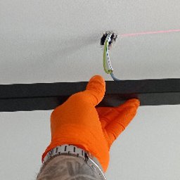 Jeremiasz Rogóż ELEKTRA SERWIS - Instalator w pomarańczowej rękawicy przymierza czarną listwę oświetleniową z reflektorami do sufitu, widoczna wiązka lasera wyznaczająca linię montażu.