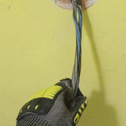 Jeremiasz Rogóż ELEKTRA SERWIS - Elektryk w rękawicy ochronnej wkłada kabel zasilający do otworu w żółtej ścianie, widoczne uszkodzenia tynku wokół otworu.