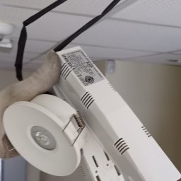 Jeremiasz Rogóż ELEKTRA SERWIS - Biała oprawa LED z zasilaczem w dłoni w rękawicy roboczej, widoczna na tle sufitu podwieszanego z czarnym kablem mocującym.