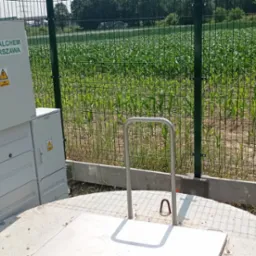 Dwie szare skrzynki elektryczne z logo Metalchem Warszawa i ostrzeżeniem o wysokim napięciu, umieszczone obok betonowej studzienki z metalowym uchwytem, całość ogrodzona zieloną siatką na tle pola...