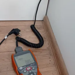 Pomarańczowy miernik Sonel MZC-304 wyświetlający parametry elektryczne, podłączony przewodem do gniazdka z włożoną wtyczką, ustawiony na podłodze z paneli.