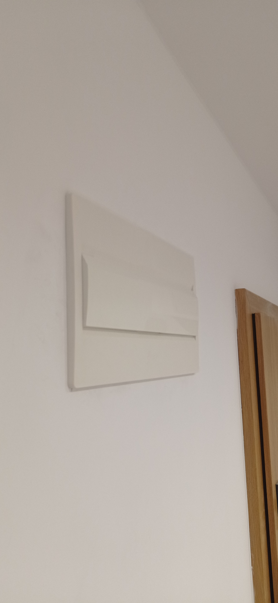 Biała tablica rozdzielcza z bezpiecznikami zamontowana na białej ścianie, widoczny fragment drewnianych drzwi po prawej stronie.