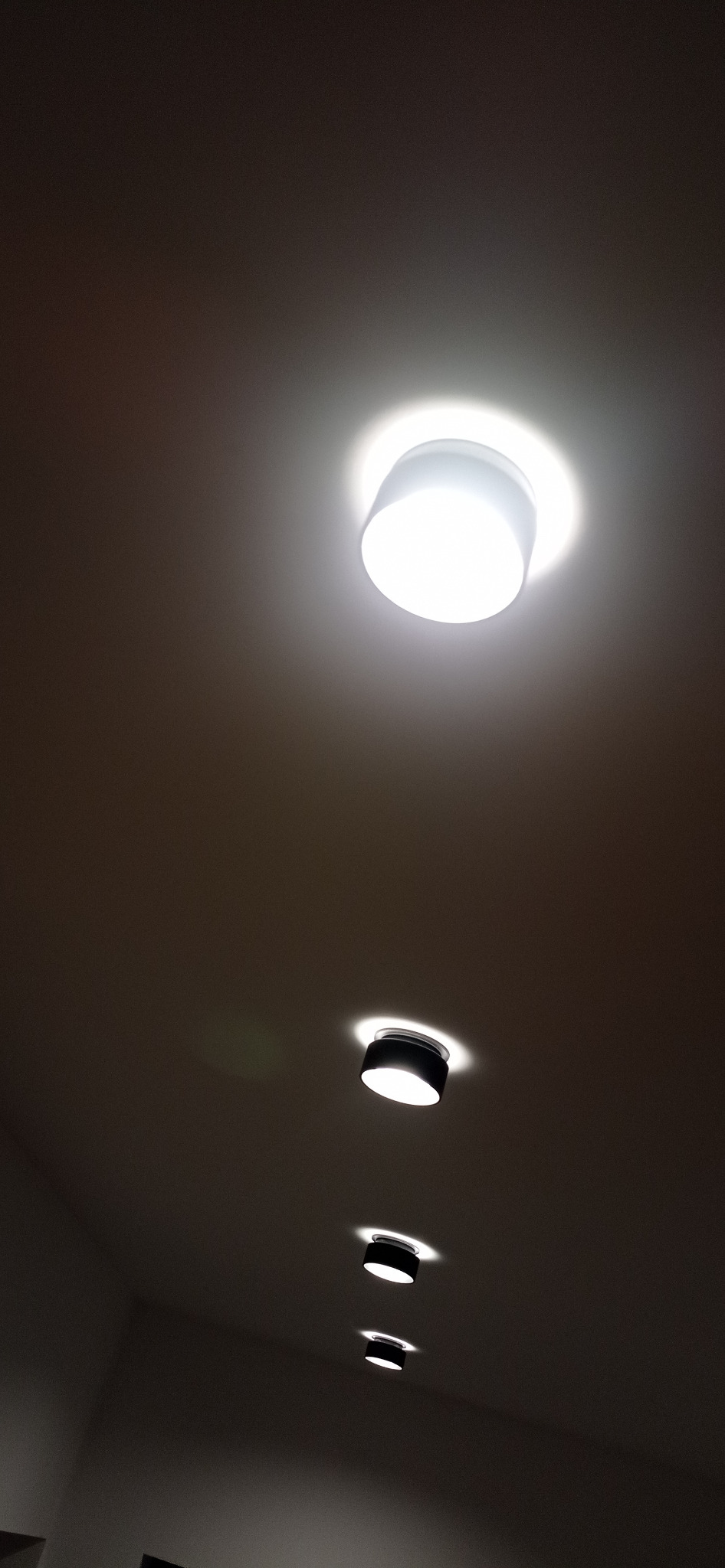 Równomiernie rozmieszczone, okrągłe lampy sufitowe LED w rzędzie, oświetlające ciemne pomieszczenie, z dominującym światłem od pierwszej lampy.