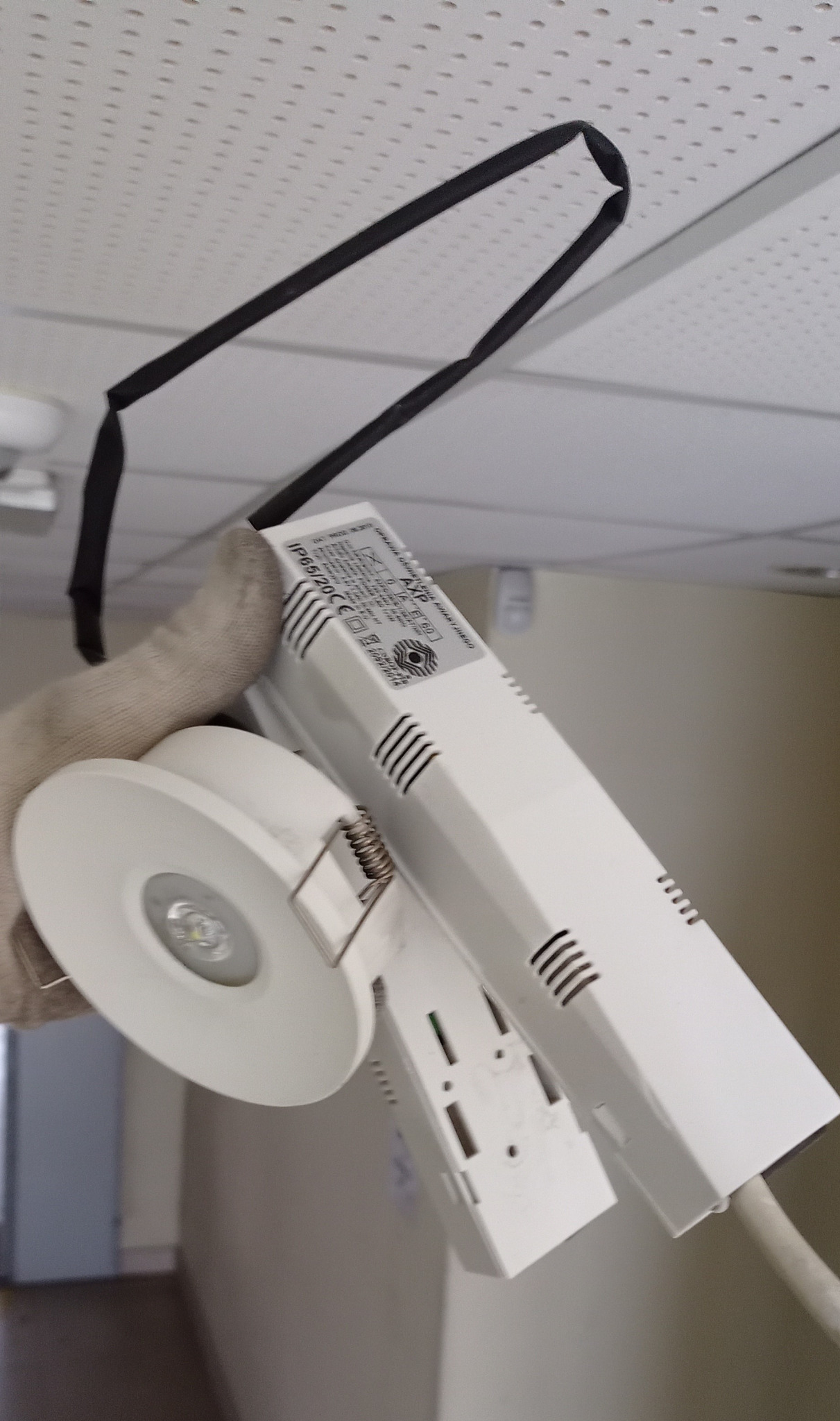 Biała oprawa LED z zasilaczem w dłoni w rękawicy roboczej, widoczna na tle sufitu podwieszanego z czarnym kablem mocującym.