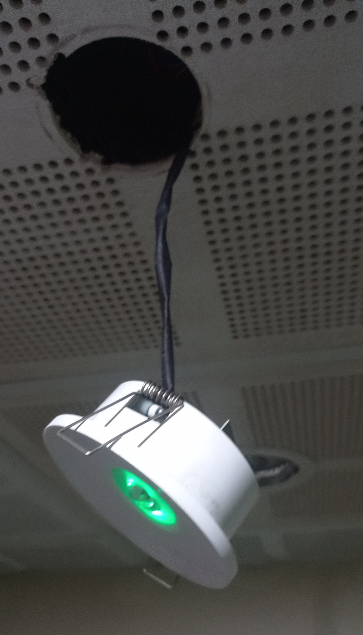 Biała oprawa oświetleniowa LED z zielonym światłem zamontowana w suficie podwieszanym, widoczny kabel zasilający i otwór w suficie.