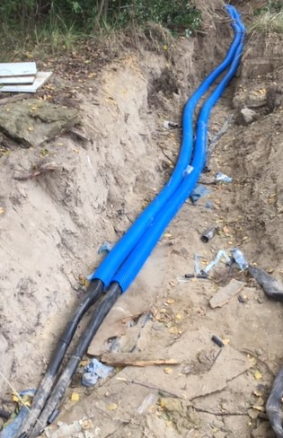 Dwa niebieskie karbowane przewody ułożone w wykopie ziemnym, z widocznymi czarnymi kablami wewnątrz, częściowo zasypane ziemią.