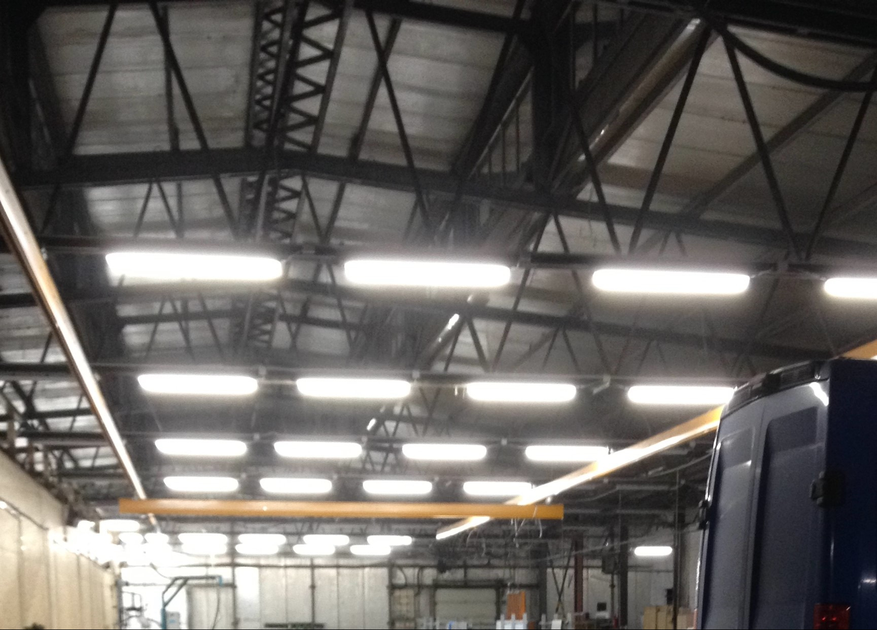 Oświetlenie hali przemysłowej z rzędami lamp LED zamontowanych pod ciemną, stalową konstrukcją dachu, widoczny fragment zaparkowanego busa.