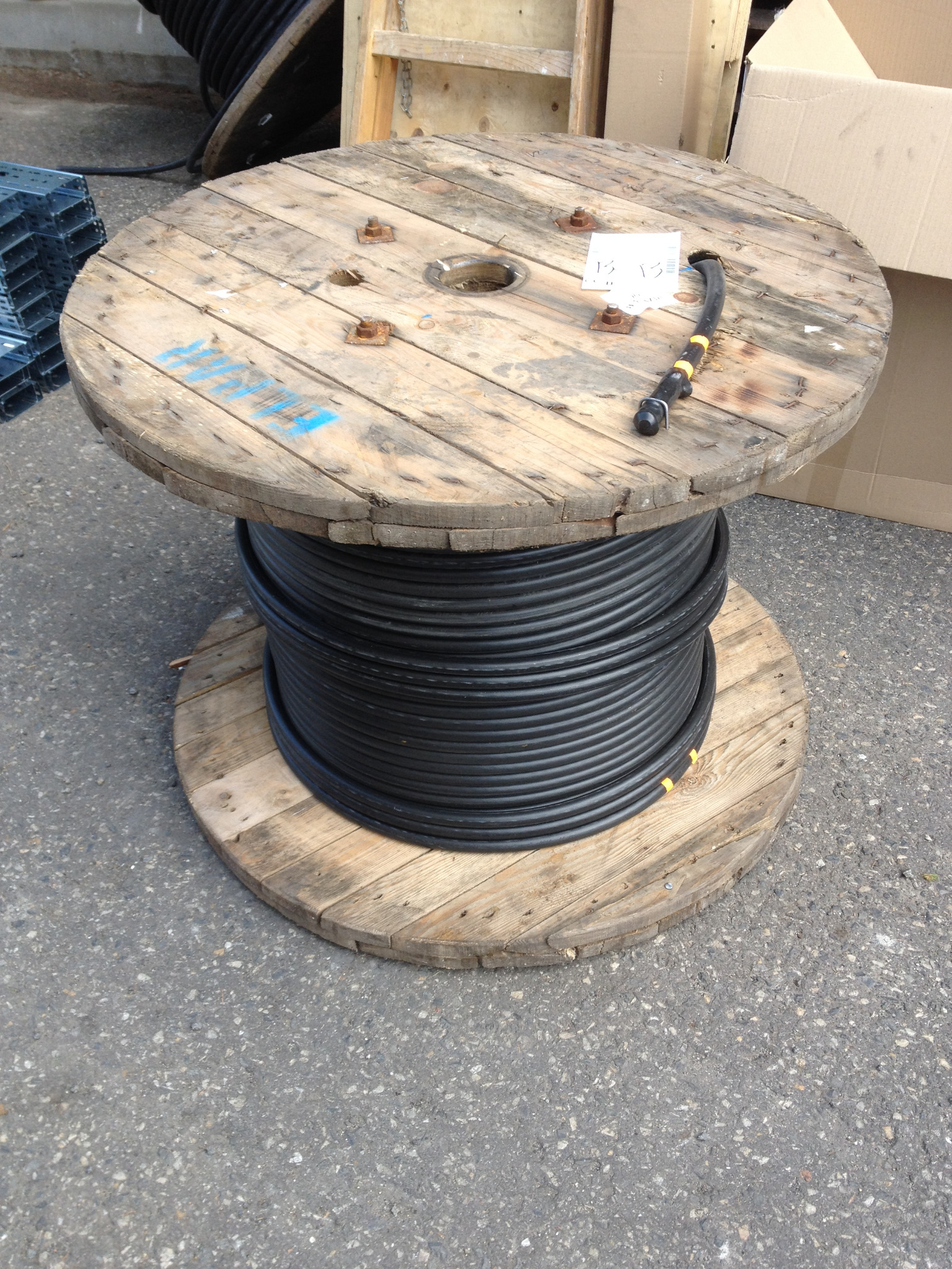 Drewniana szpula z nawiniętym czarnym kablem elektrycznym, widoczny fragment przewodu z żółtymi oznaczeniami, szpula stoi na szarym asfalcie.