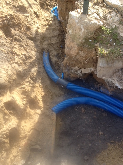 Wykop w ziemi z ułożonymi niebieskimi rurami karbowanymi, prawdopodobnie do instalacji elektrycznej pod ziemią, widoczne kamienie i piasek