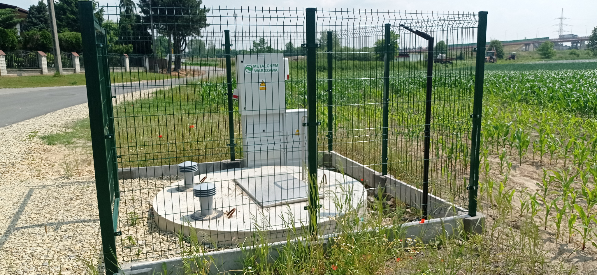 Zewnętrzna szafka elektryczna z logo Metalchem Warszawa, otoczona metalową siatką, na tle pola kukurydzy i wiaduktu.