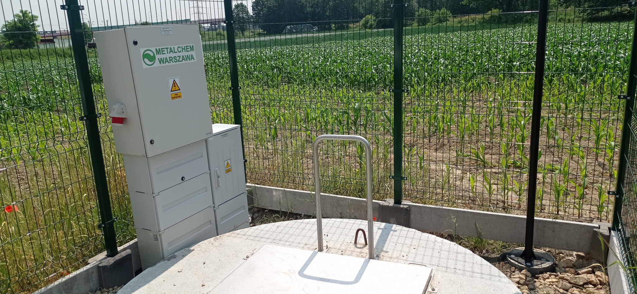 Dwie szare skrzynki elektryczne z logo Metalchem Warszawa i ostrzeżeniem o wysokim napięciu, umieszczone obok betonowej studzienki z metalowym uchwytem, całość ogrodzona zieloną siatką na tle pola...