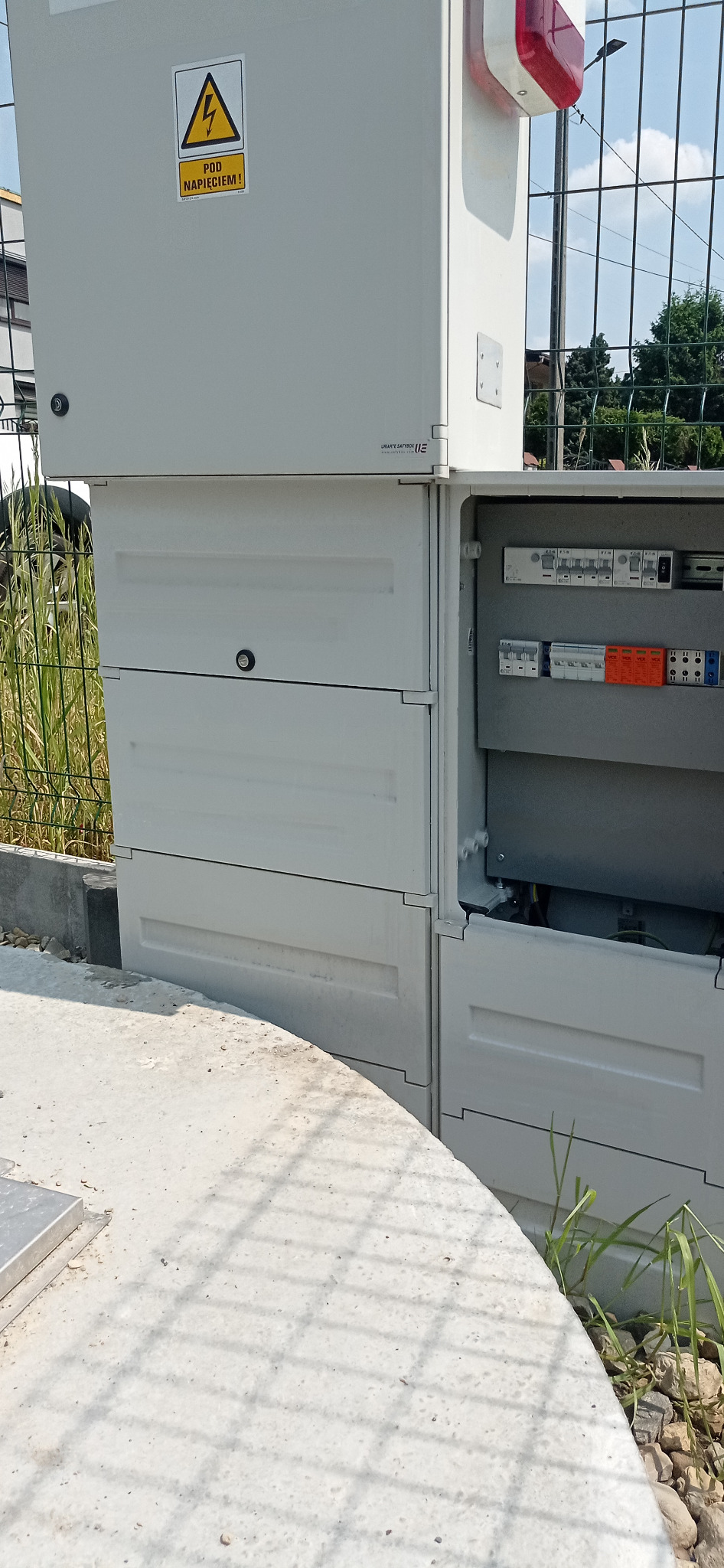 Szara, modułowa skrzynka elektryczna z otwartymi drzwiczkami, ukazująca wewnętrzne okablowanie i zabezpieczenia, umieszczona na betonowej podstawie, z widocznym znakiem ostrzegawczym o wysokim...