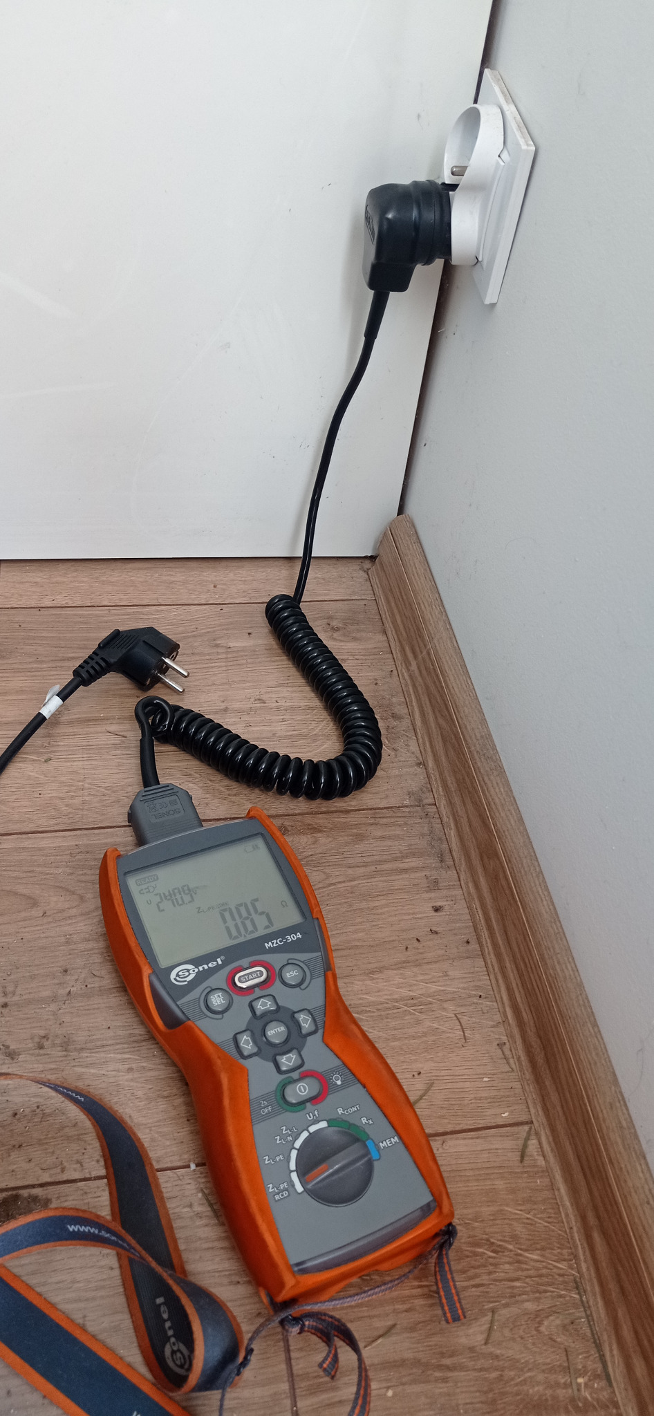 Pomarańczowy miernik Sonel MZC-304 wyświetlający parametry elektryczne, podłączony przewodem do gniazdka z włożoną wtyczką, ustawiony na podłodze z paneli.