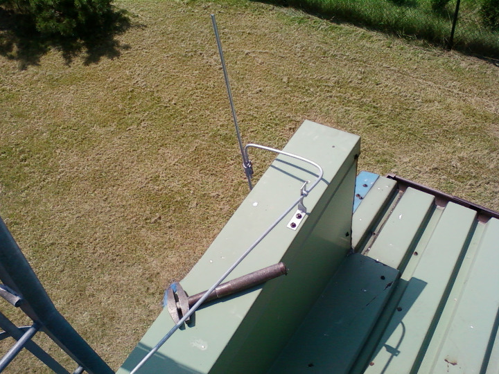 Metalowy maszt odgromowy zamontowany na zielonym dachu z blachy trapezowej, widok z góry na tle trawnika.