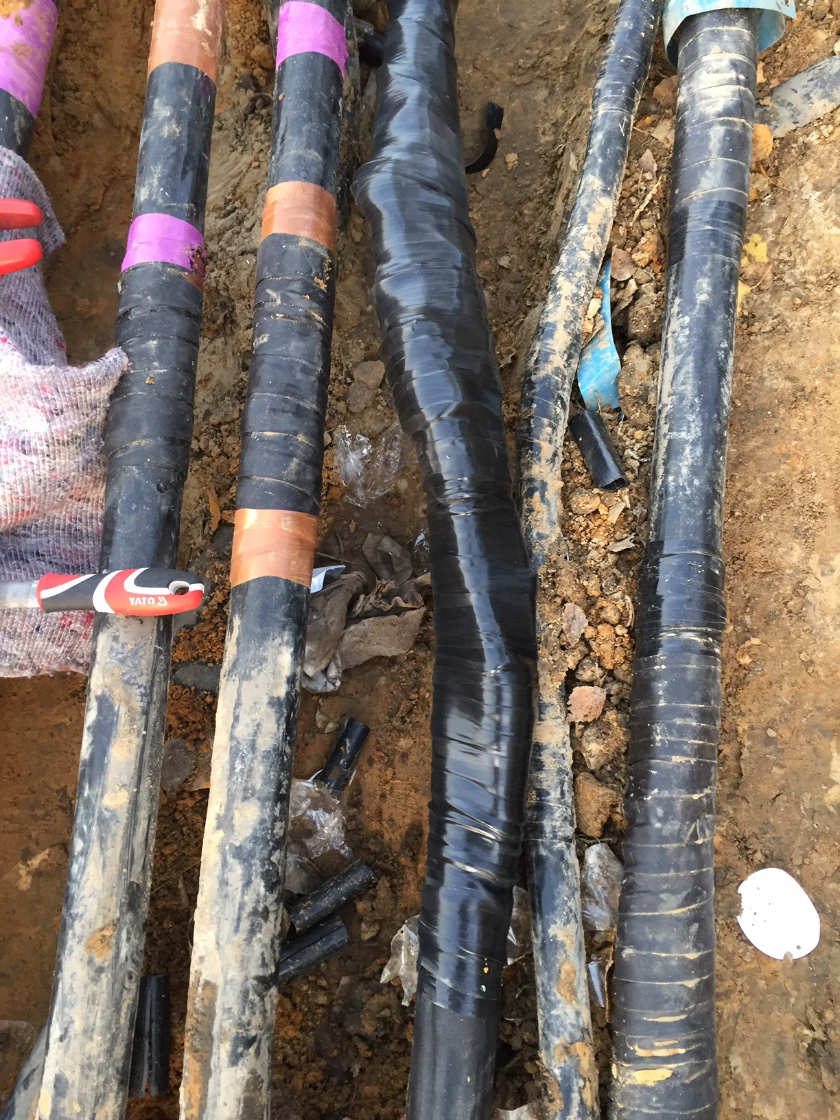 Kable elektryczne w wykopie, zabezpieczone taśmą izolacyjną w różnych kolorach, widoczny nóż monterski Yato i resztki folii ochronnej.