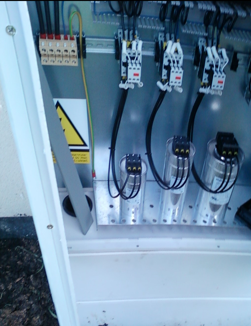 Wnętrze rozdzielnicy elektrycznej z kondensatorami, stycznikami i listwami zaciskowymi, widoczny znak ostrzegawczy, ujęcie z dołu.