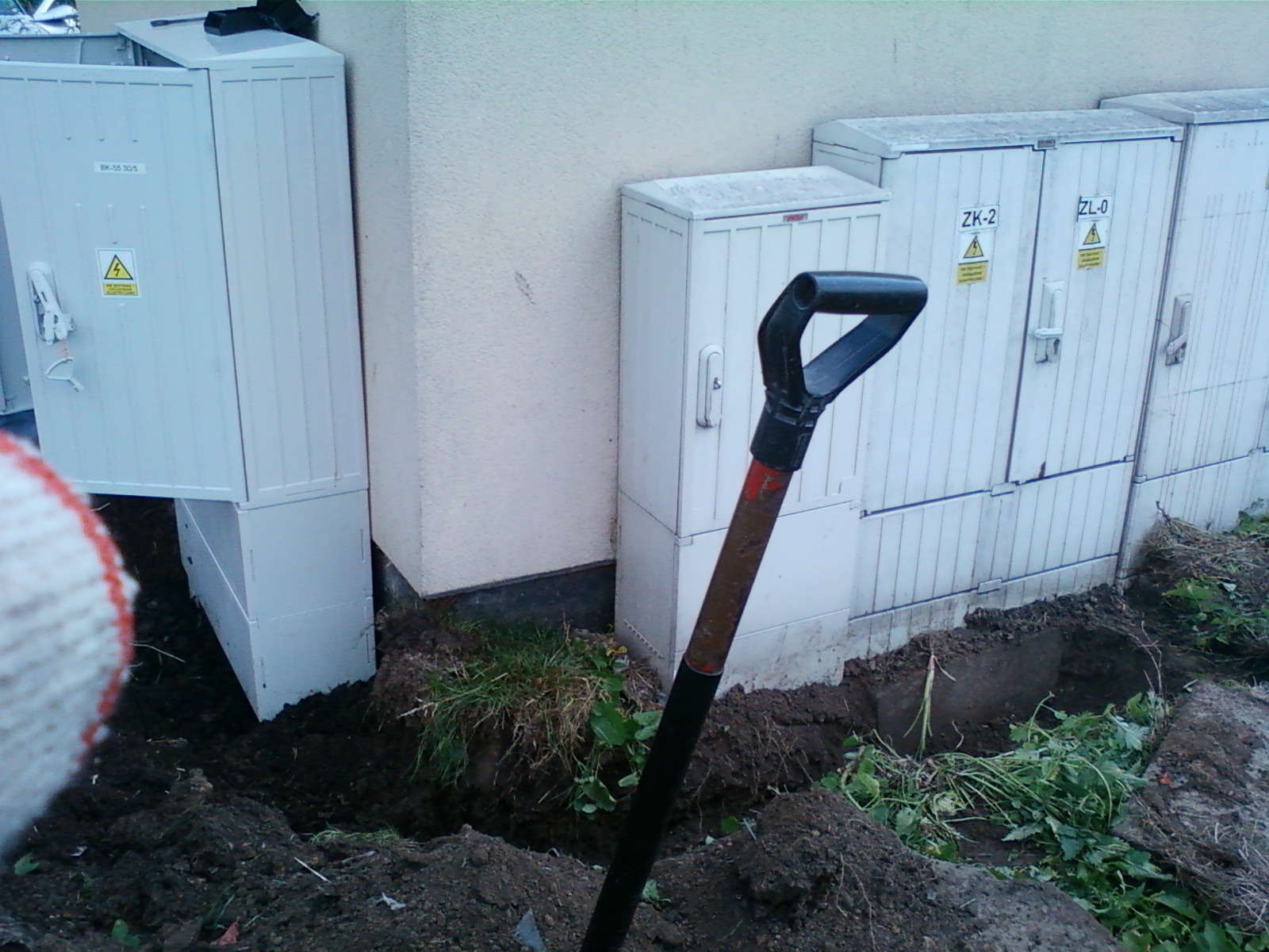 Prace ziemne przy instalacji elektrycznej: widoczne skrzynki elektryczne z ostrzeżeniami, wykopany rów i łopata z czarną rączką w ziemi, fragment rękawicy roboczej w lewym dolnym rogu.