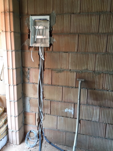 Otwarta puszka elektryczna wmurowana w ścianę z czerwonej cegły, z wystającymi białymi kablami. Część kabli poprowadzona w dół ściany w wyżłobieniu. Obok ściany oparta łopata.