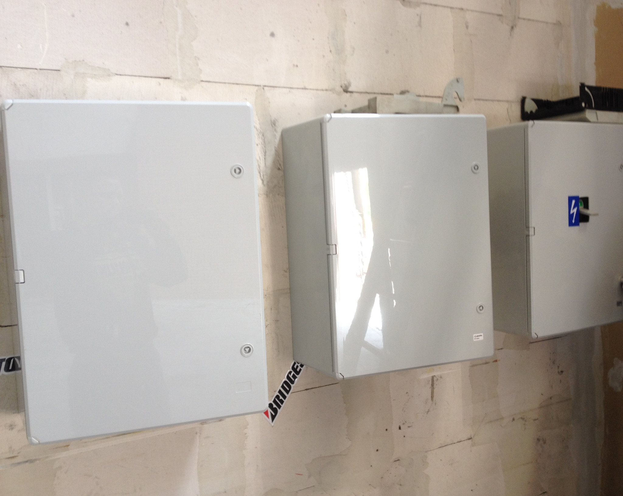 Trzy szare skrzynki elektryczne zamontowane na surowej ścianie z widocznymi śladami po remoncie; na jednej ze skrzynek naklejka z symbolem błyskawicy i przełącznik.