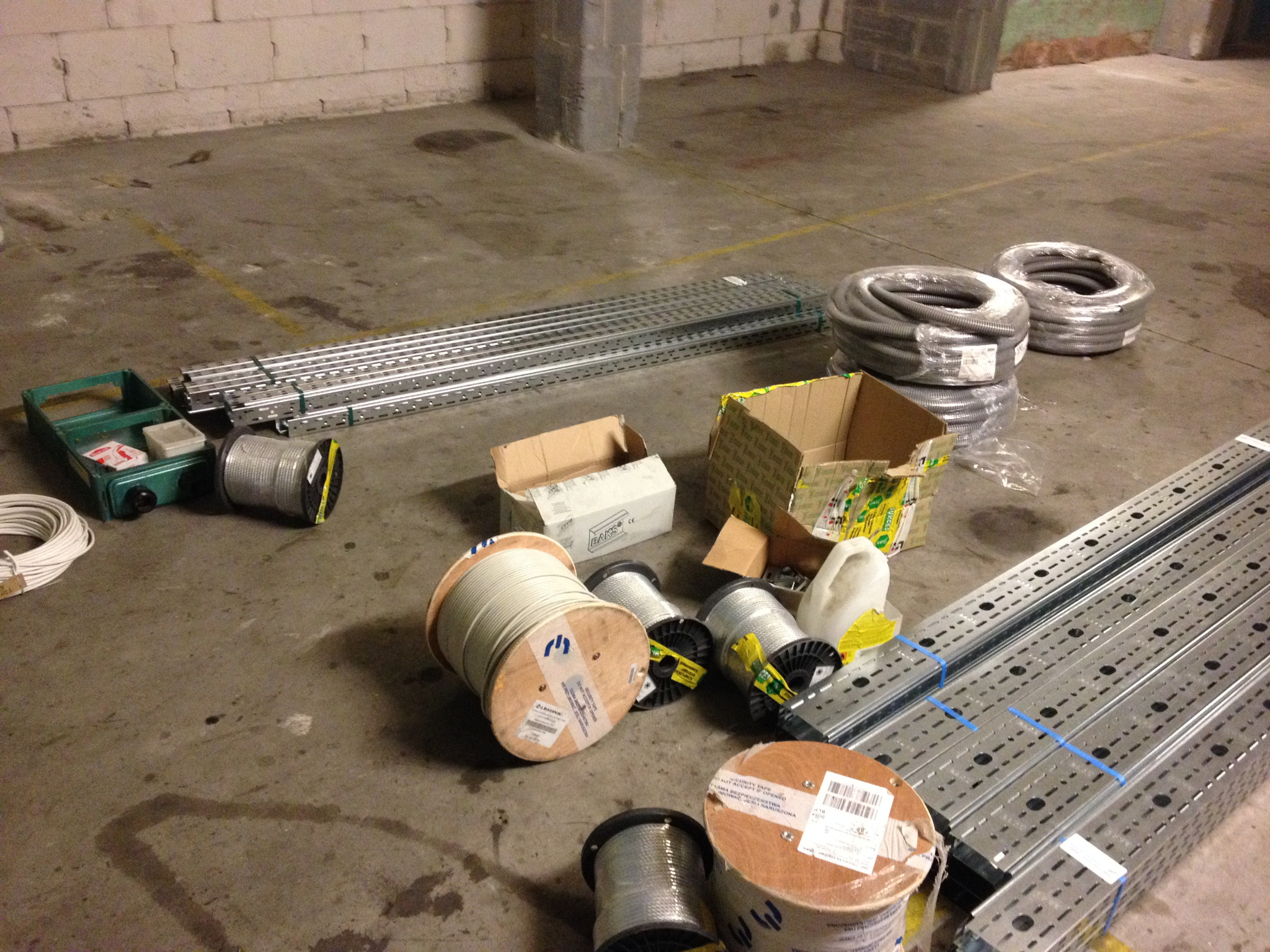 Spiętrzone rolki kabli elektrycznych, metalowe korytka kablowe i kartony na betonowej podłodze w magazynie.