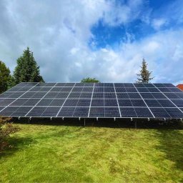 INOWA SPÓŁKA Z OGRANICZONĄ ODPOWIEDZIALNOŚCIĄ - Instalacja fotowoltaiczna na zielonym wzgórzu, panele ustawione na metalowej konstrukcji, błękitne niebo z chmurami w tle, widok z dołu.