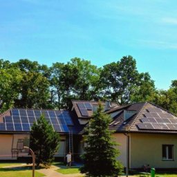 INOWA SPÓŁKA Z OGRANICZONĄ ODPOWIEDZIALNOŚCIĄ - Dom jednorodzinny z panelami fotowoltaicznymi na dachu, otoczony zielenią. Widoczna drabina oparta o dach i sylwetka osoby w pobliżu.