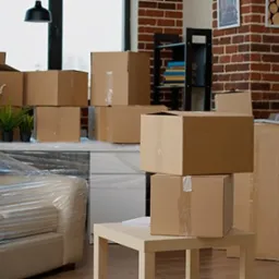 Pokój przygotowany do transportu: sofa owinięta folią, stos kartonowych pudeł, drabina oparta o ścianę z cegły, rośliny doniczkowe na meblach.