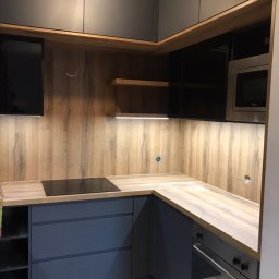 Studio Mebli Twoja Kuchnia.EU - Szara narożna zabudowa kuchenna z drewnianym blatem i panelem ściennym, oświetleniem LED podszafkowym oraz wbudowanym sprzętem AGD, takim jak płyta indukcyjna i mikrofalówka.