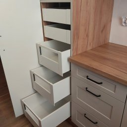 Studio Mebli Twoja Kuchnia.EU - Wnętrze szafy z wysuwanymi, białymi szufladami o różnych głębokościach, widoczne zawiasy drzwi i fragment blatu drewnianego.