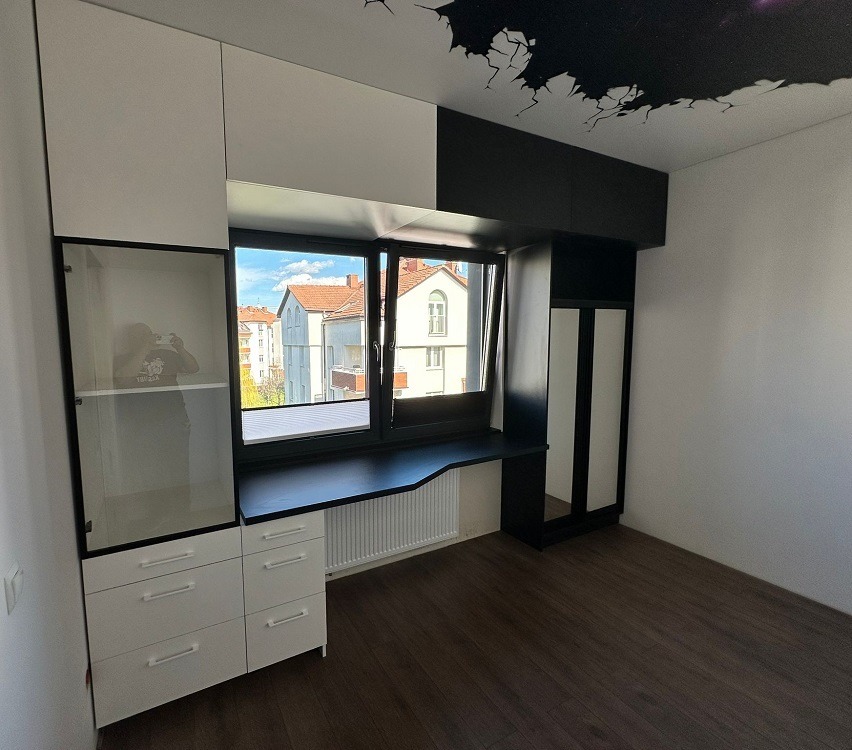 Biało-czarna zabudowa meblowa z szafkami, szufladami i przeszkloną witryną, wkomponowana w przestrzeń okienną z blatem roboczym, uzupełniona o szafę z lustrem i czarną dekoracją sufitową.