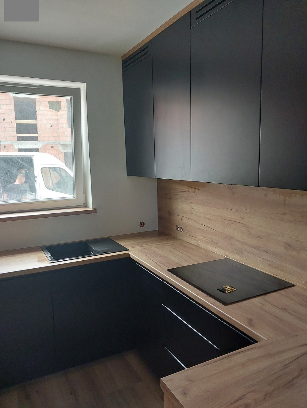 Nowoczesna kuchnia z czarnymi frontami szafek i drewnianym blatem, widok na okno z budową w tle.