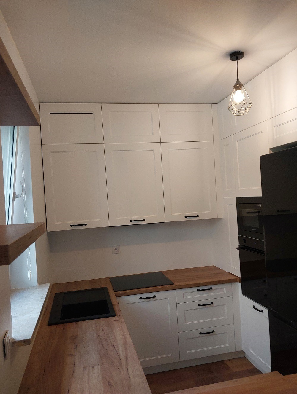 Biała zabudowa kuchenna z drewnianym blatem, czarnymi uchwytami i płytą indukcyjną, oświetlona lampą wiszącą w stylu industrialnym.