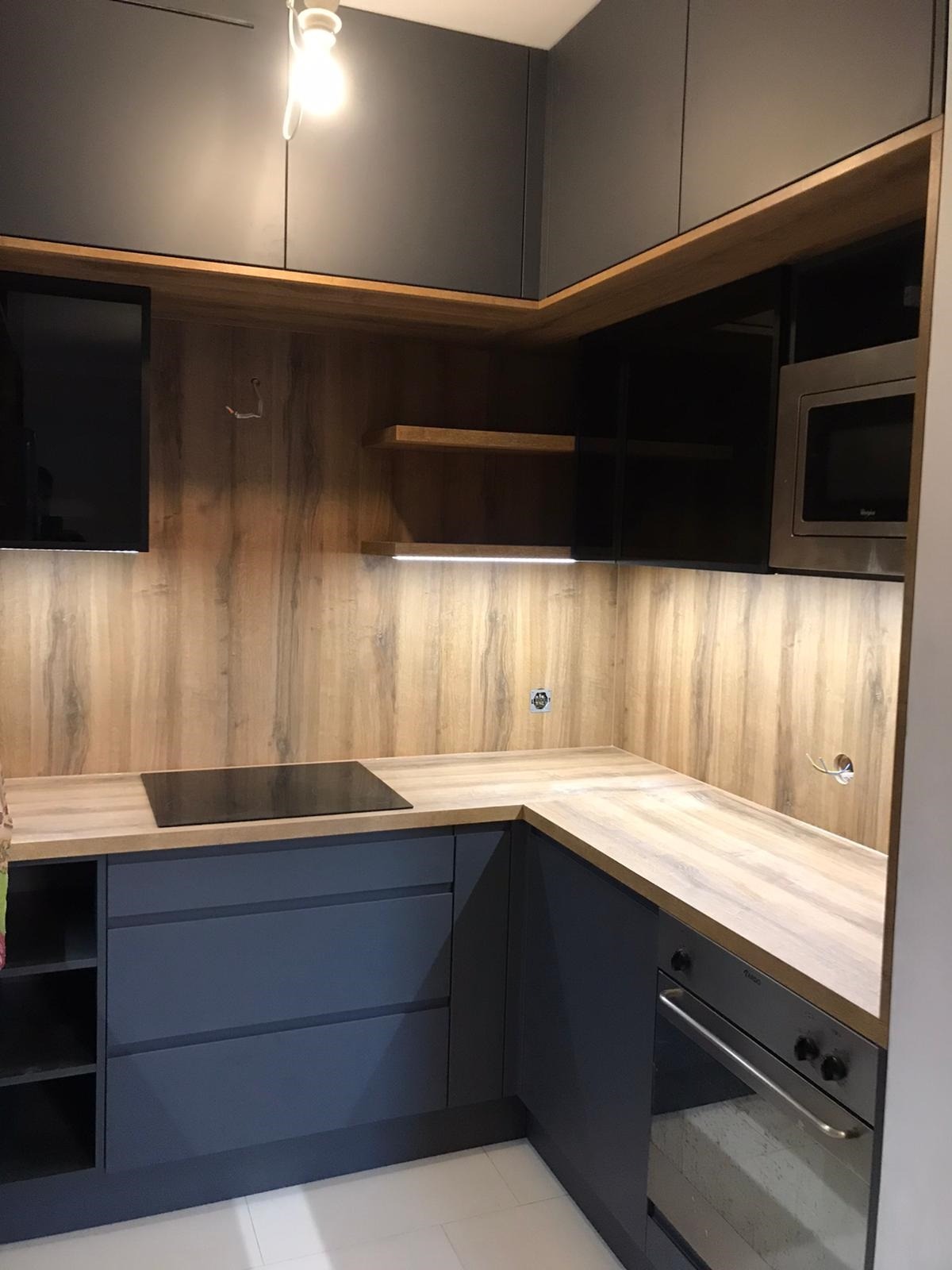 Szara narożna zabudowa kuchenna z drewnianym blatem i panelem ściennym, oświetleniem LED podszafkowym oraz wbudowanym sprzętem AGD, takim jak płyta indukcyjna i mikrofalówka.