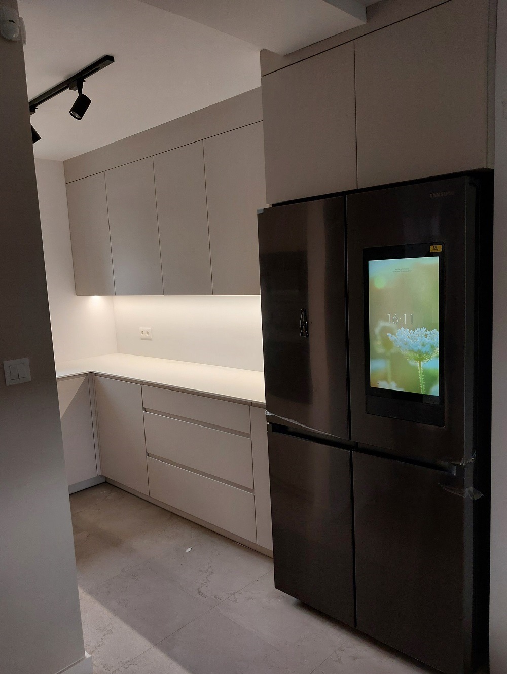 Nowoczesna kuchnia z gładkimi, jasnymi frontami szafek i dużym, czarnym lodówko-zamrażarką Samsung z ekranem dotykowym wkomponowana w zabudowę.