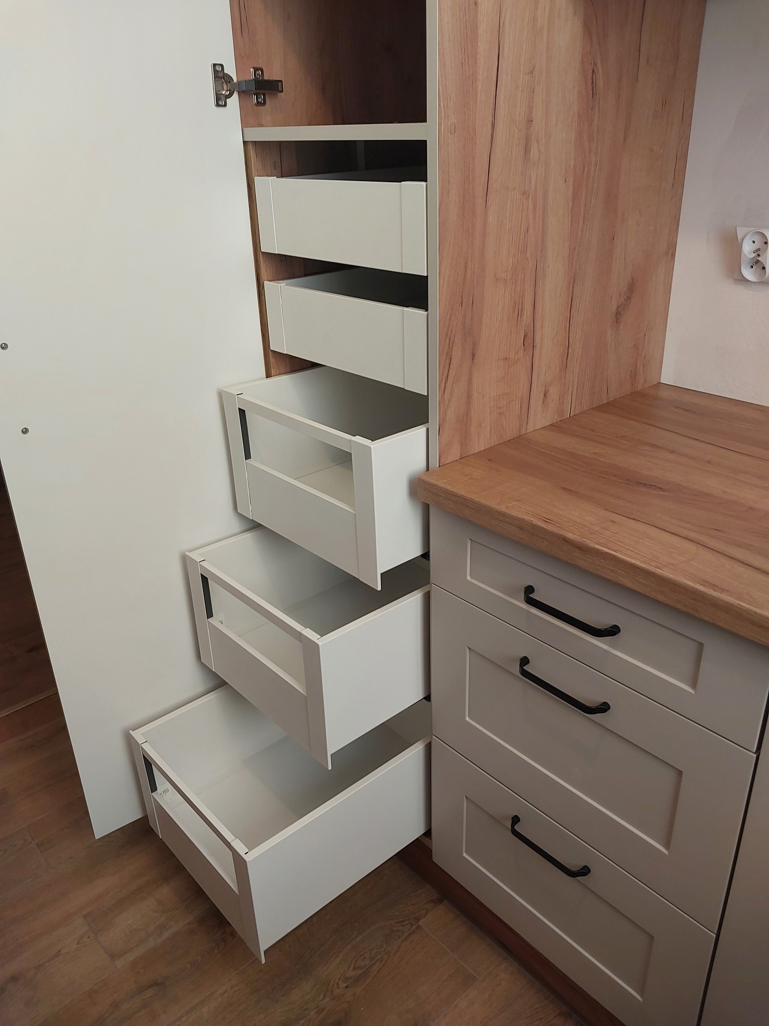 Wnętrze szafy z wysuwanymi, białymi szufladami o różnych głębokościach, widoczne zawiasy drzwi i fragment blatu drewnianego.