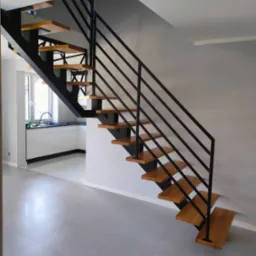 Nowoczesne schody o konstrukcji stalowej i drewnianych stopniach, z minimalistyczną balustradą z czarnych prętów, w jasnym wnętrzu z widoczną kuchnią w tle.