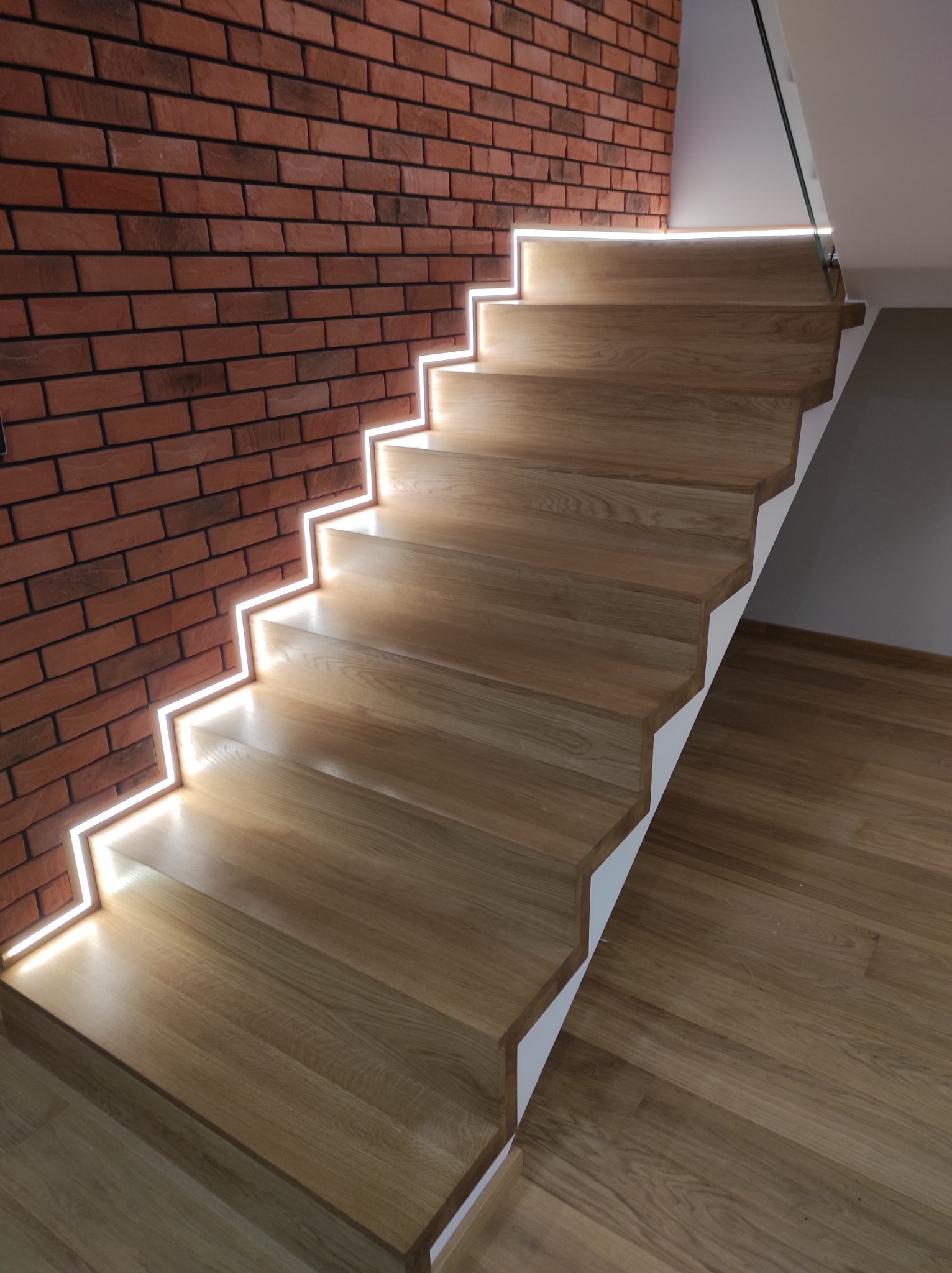 Nowoczesne schody z jasnego drewna z podświetleniem LED wzdłuż stopni, kontrastujące z czerwoną ścianą z cegły i drewnianą podłogą.