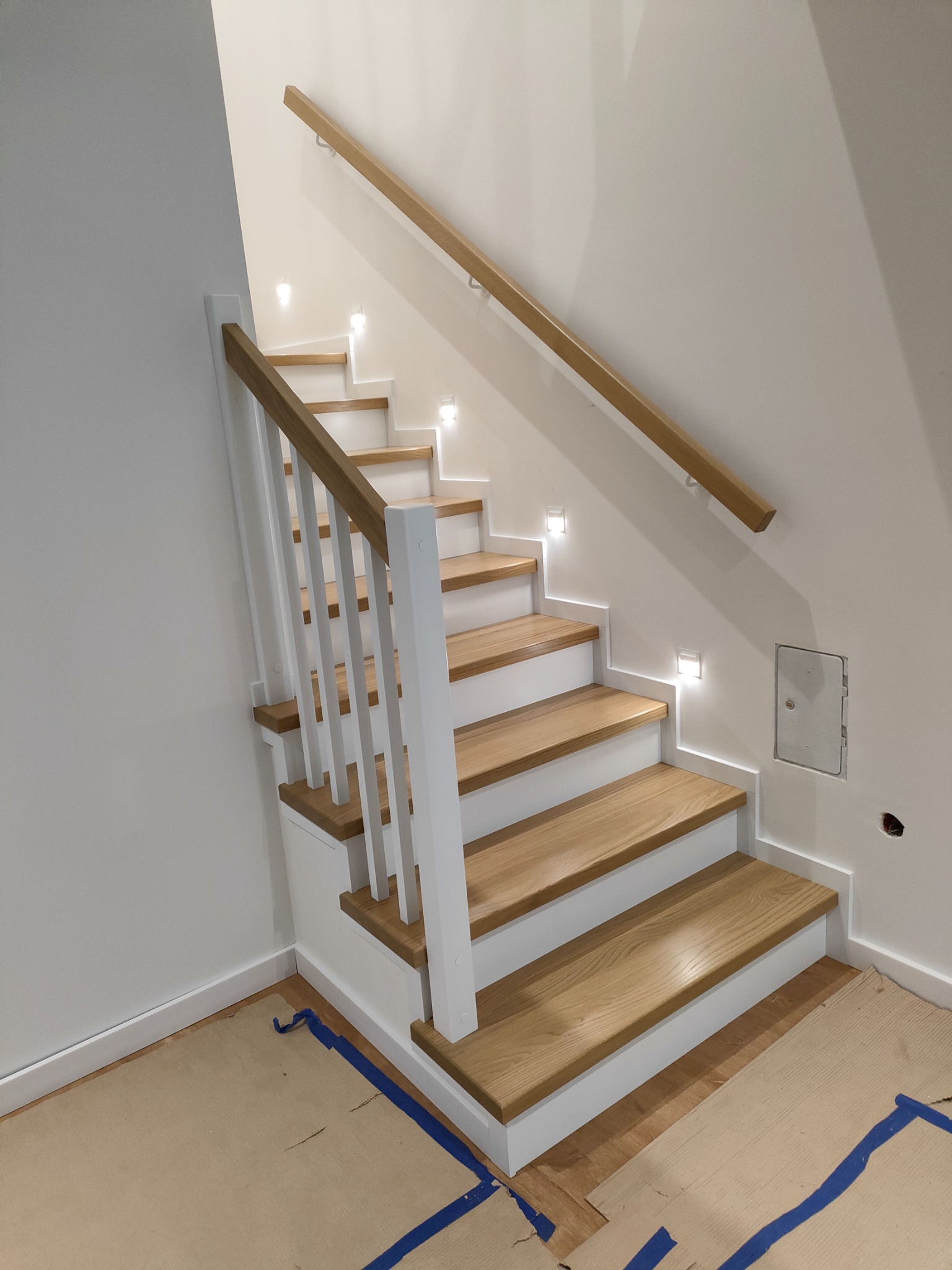 Nowoczesne schody drewniane z białymi podstopnicami i drewnianymi stopniami, oświetlone małymi lampkami w ścianie, zabezpieczone kartonem i taśmą malarską na podłodze.