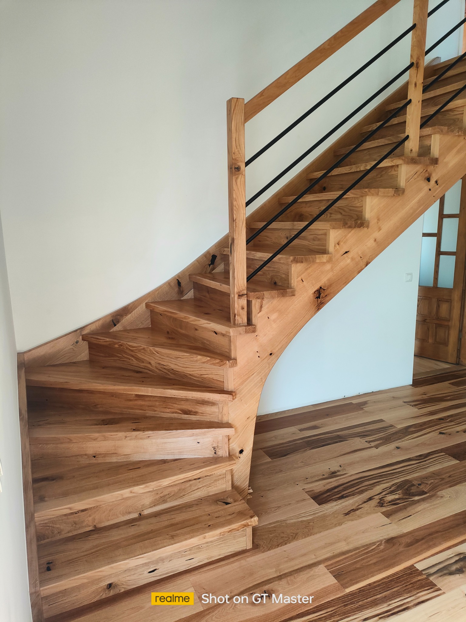 Jasne drewniane schody z czarnymi metalowymi balustradami, widoczne sęki i unikalny wzór słojów drewna, dopasowane drewniane wykończenie podłogi, w tle drzwi.