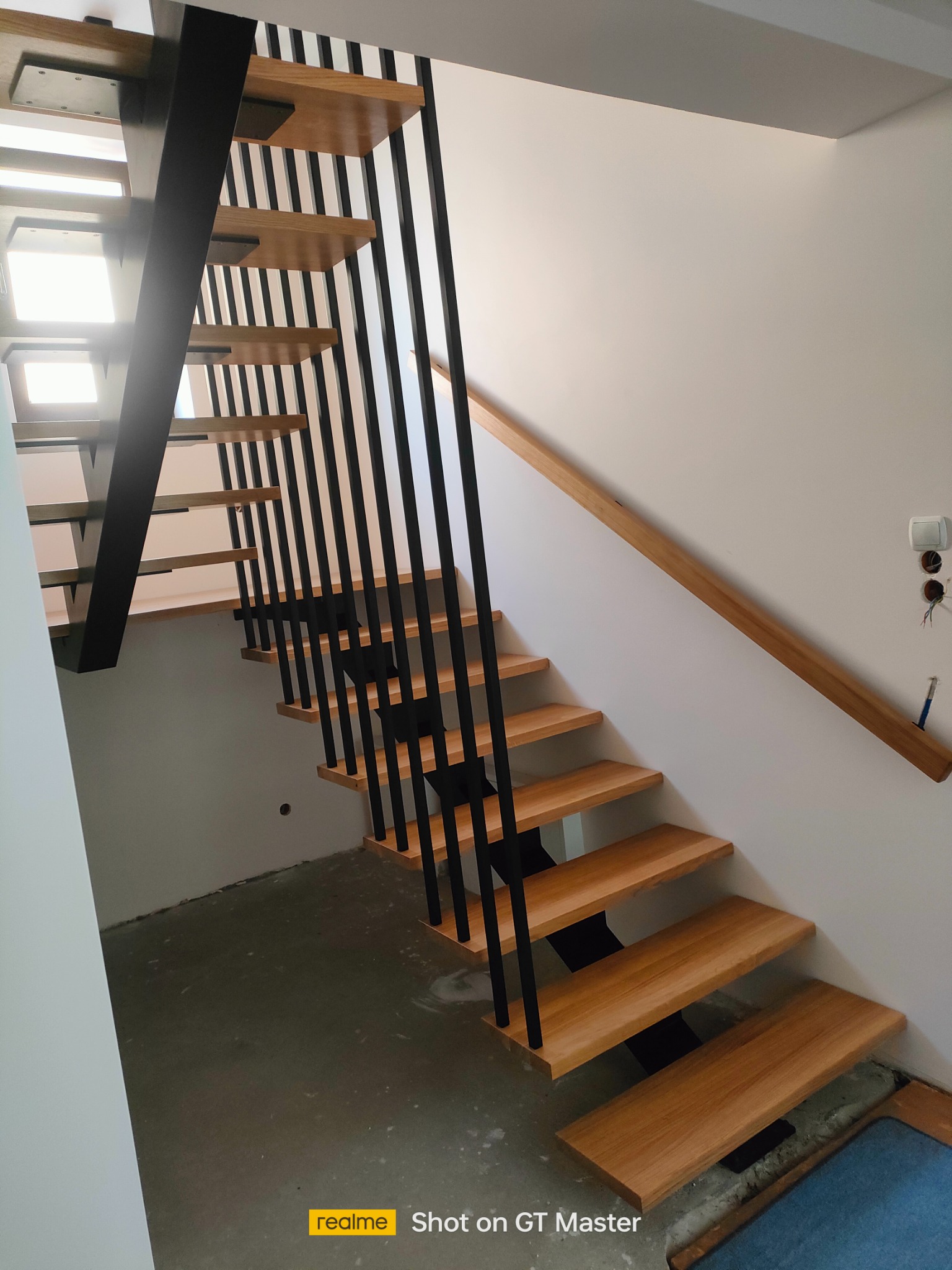 Nowoczesne schody z drewnianymi stopniami i czarnymi metalowymi balustradami w jasnym wnętrzu, widoczne przygotowania do wykończenia podłogi.