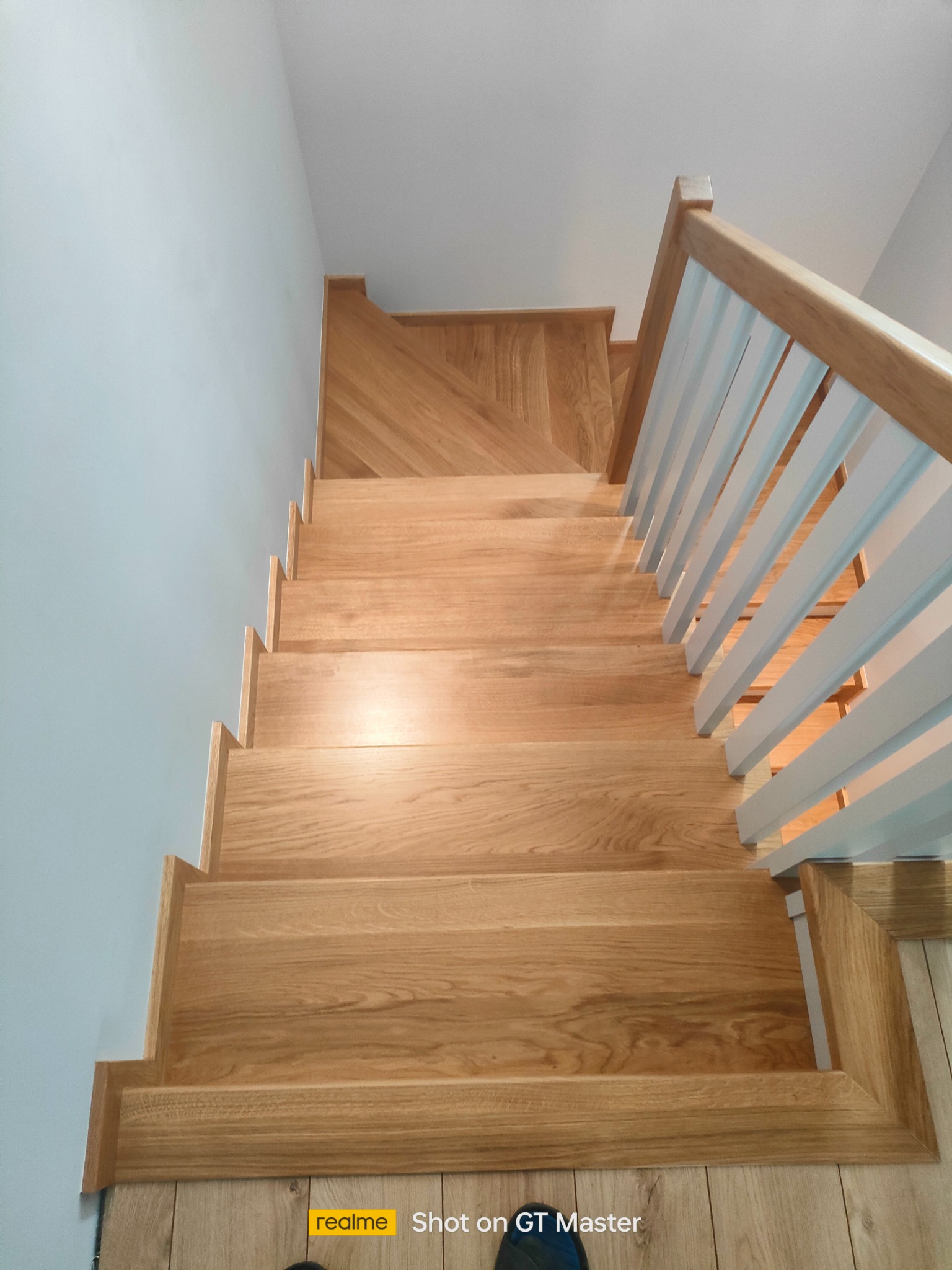 Kątowe ujęcie z góry na jasne, drewniane schody z białymi, pionowymi balustradami, prowadzące w dół, z drewnianą podłogą na dole i szarą ścianą po lewej.